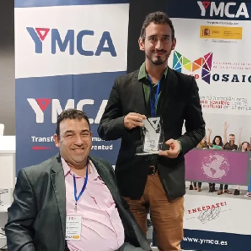 Una persona de pie sostiene un trofeo junto a otra sentada, ambos en un evento con pancartas que dicen “YMCA” y “MOSAICO” de fondo.