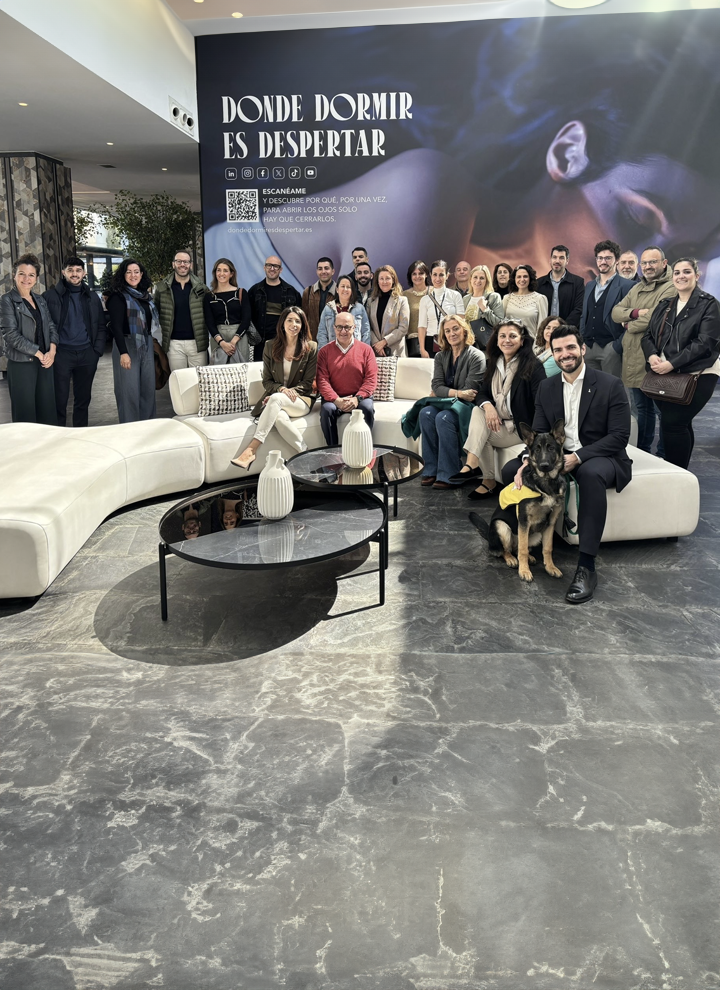 En la imagen se ve al personal de Paradores de Turismo de España junto con personal trabajador del hotel y Lía, la perra de ILUNION Atrium, en el hall del hotel frente a la nueva campaña 