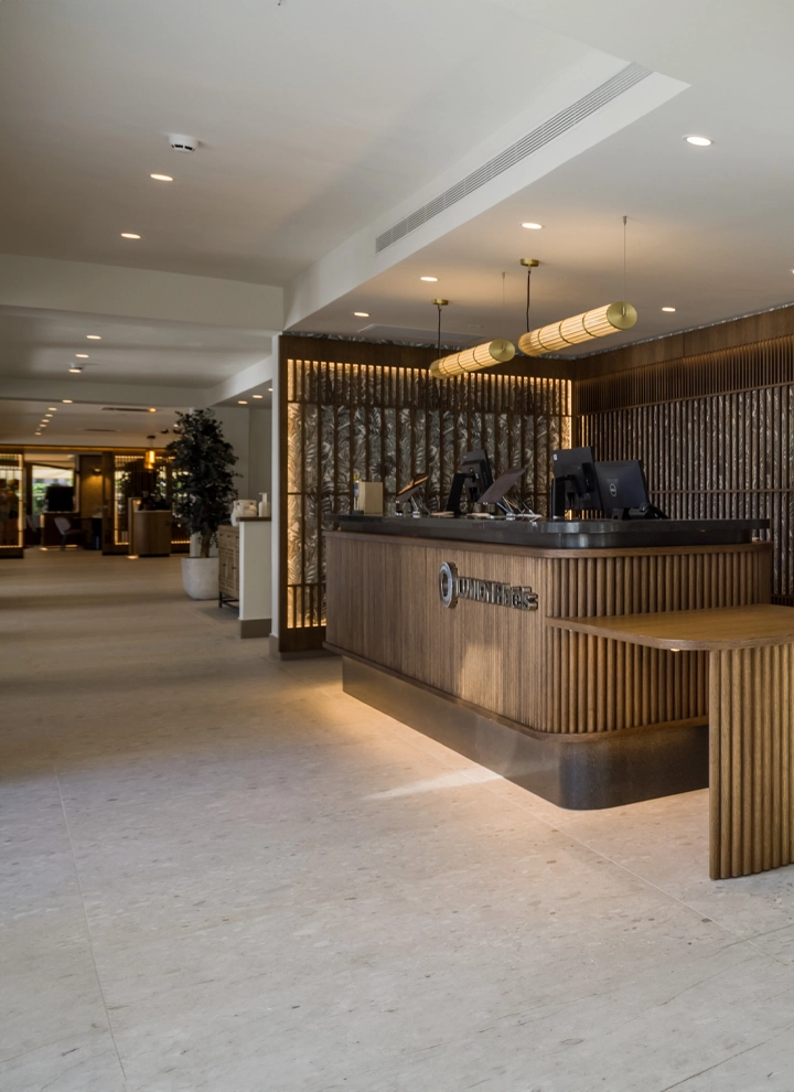 La imagen muestra una elegante y moderna área de recepción del renovado ILUNION Caleta Park. La recepción presenta un diseño contemporáneo con detalles cálidos y acogedores.