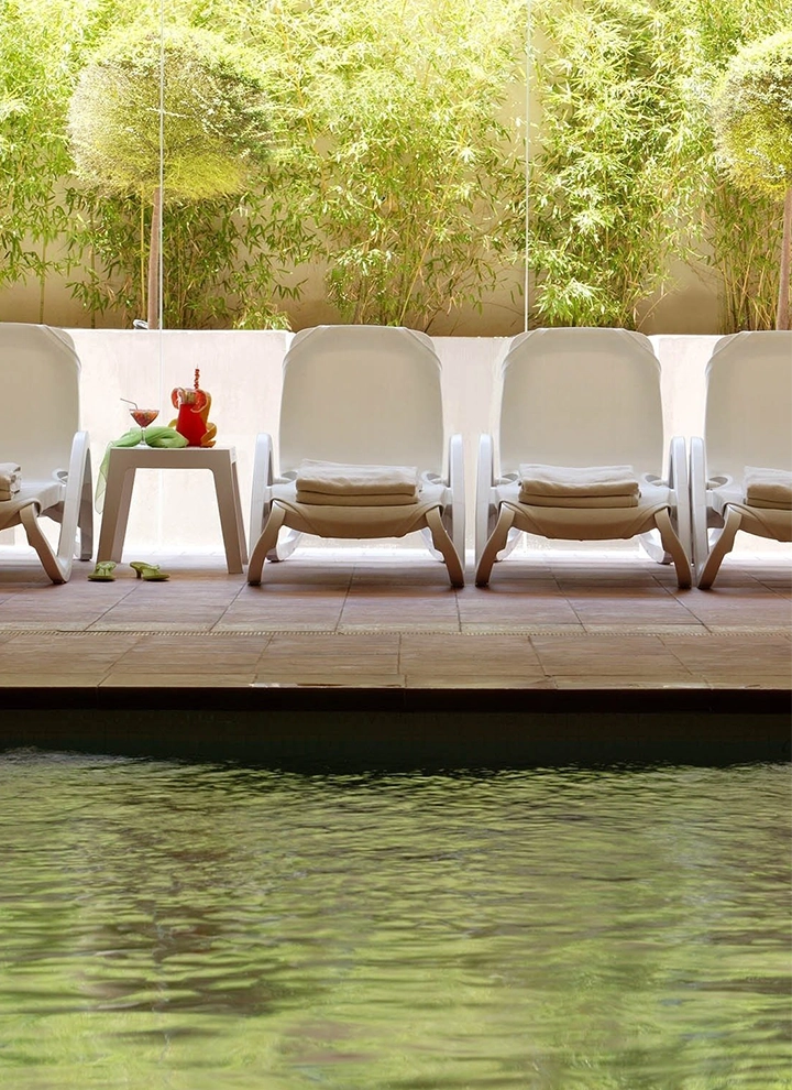 Piscina interior del hotel ILUNION Miramar. Pardes blancas y suelo de elegante losetas marrones, una piscina de agua cristalina con cuidadas y modernas hamacas blancas