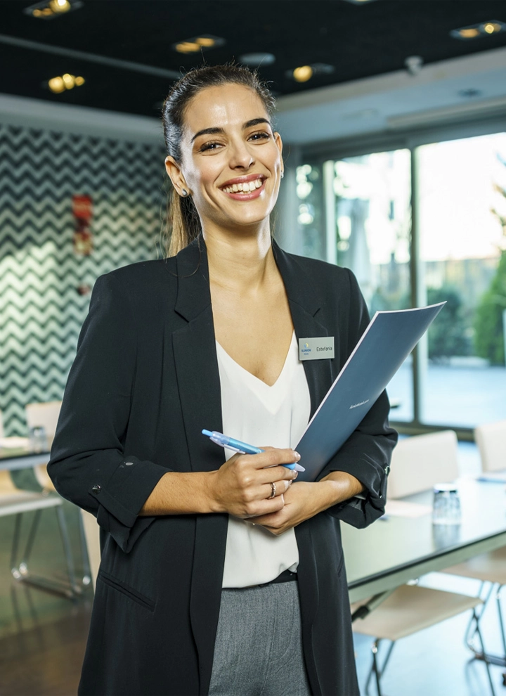 La imagen muestra a una chica sonriendo parada en el vestibulo del hotel ILUNION Atrium. La persona lleva un traje oscuro con una etiqueta de identificación y tiene los brazos cruzados.