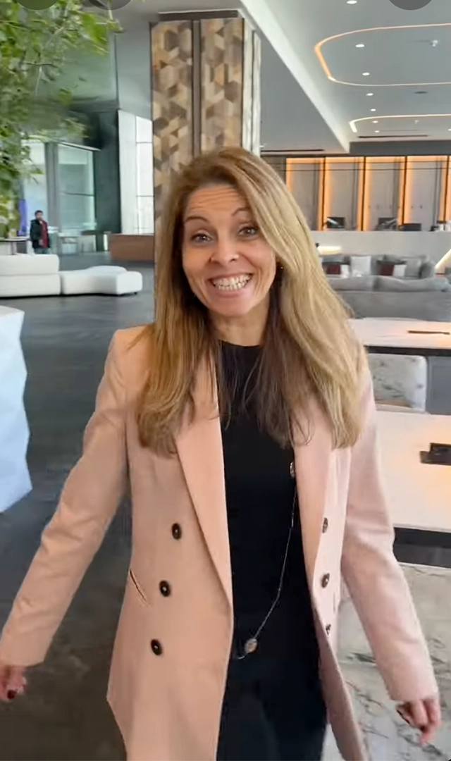 Una mujer sonriente vestida con un blazer de color rosa pálido, una camiseta negra y un collar largo, de pie en un vestíbulo con un fondo borroso.