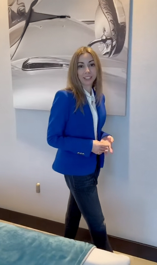 Una mujer sonriente, con un blazer azul brillante, se para en una habitación con una pintura abstracta de un coche en blanco y negro detrás de ella.