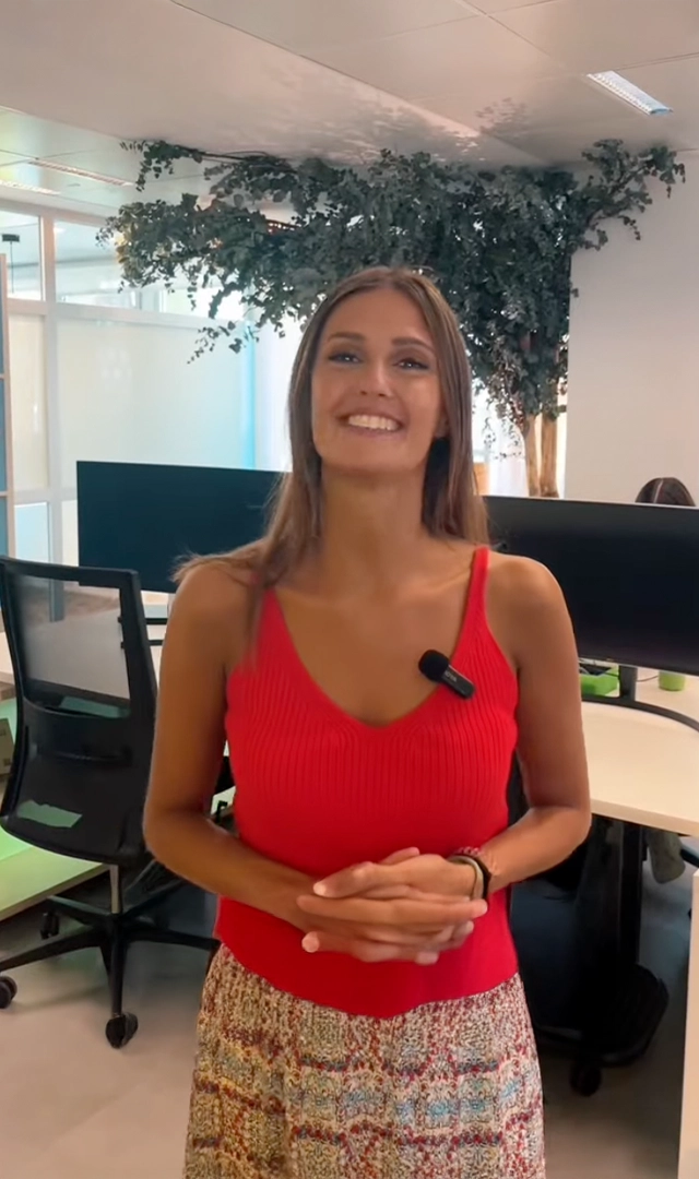 Una mujer joven, con un top rojo sin mangas y una falda estampada, sonríe a la cámara mientras se encuentra de pie en una oficina moderna y luminosa.