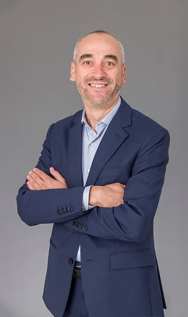Un hombre sonriendo, con traje azul marino y camisa azul claro, posa con los brazos cruzados delante de un fondo gris.