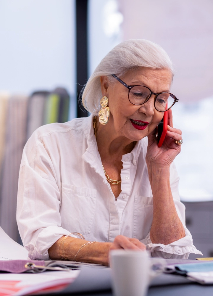 Mujer mayor sentada manteniendo una conversación telefónica, sosteniendo un smartphone con la mano izquierda mientras escribe con la mano derecha. En la pared detrás de la persona hay planos arquitectónicos fuera del foco.