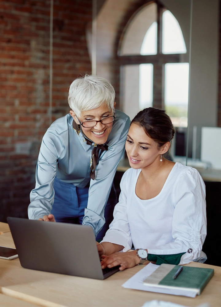 Dos personas, una de ellas sentada en una mesa con una laptop abierta frente a ellas. La otra persona parece estar de pie e inclinada ligeramente hacia la ordenador. Tiene rasgos de avanzada edad y el pelo blanco. El entorno incluye una pared de ladrillo y ventanas arqueadas, sugiriendo un ambiente de oficina o tipo loft.