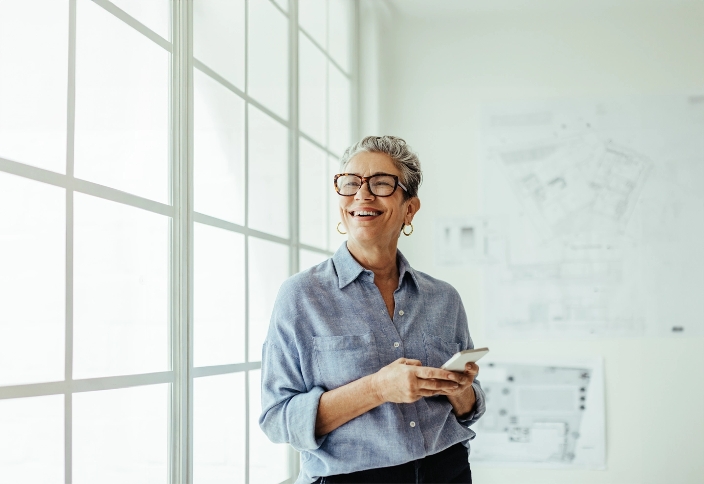 Persona de pie frente a una ventana con persianas verticales, sosteniendo un smartphone. En la pared detrás de la persona hay planos arquitectónicos visibles.