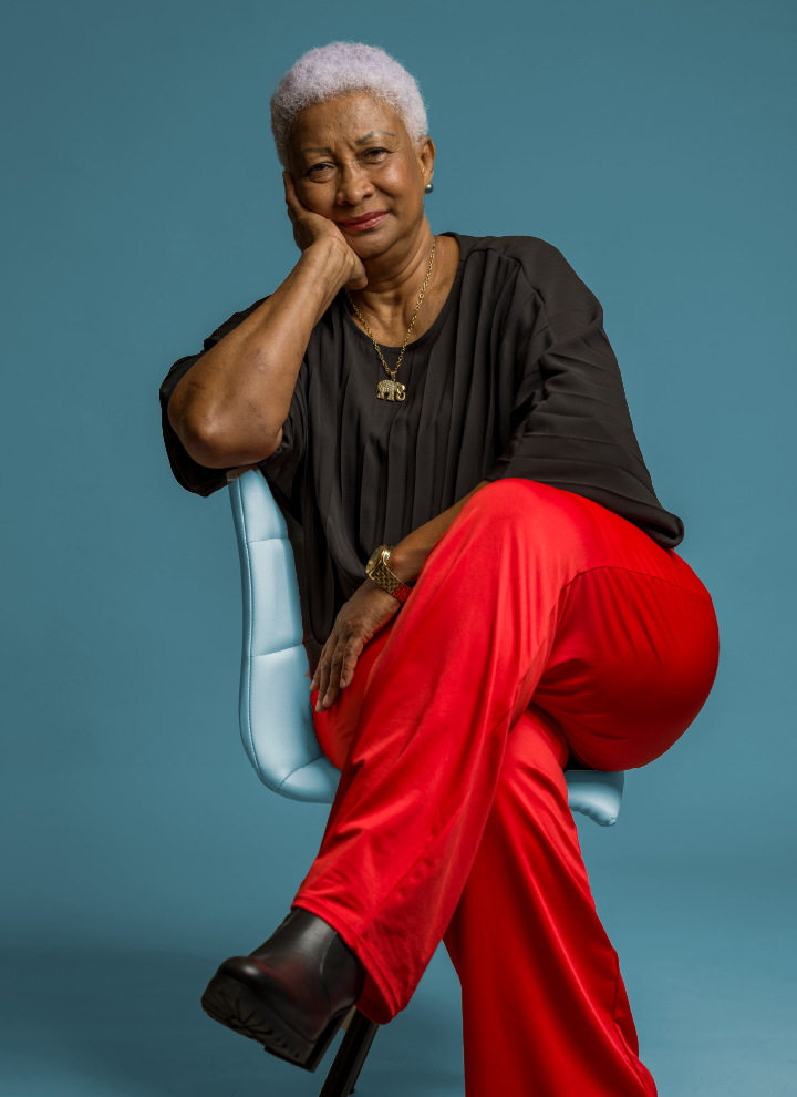 Una señora mayor con rasgos afroamericanos aparece sentada en una silla blanca y con las piernas cruzadas. Viste una blusa negra, pantalones rojos y botas negras y además tiene colgada al cuello una medalla de un elefante que está bañado en oro.