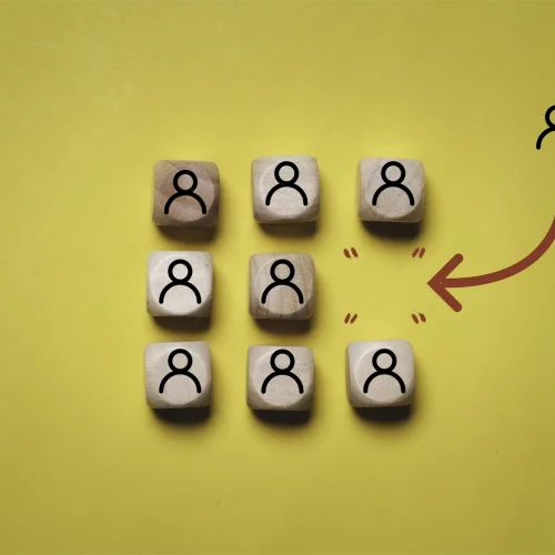 Siete bloques de madera sobre un fondo amarillo. Seis de los bloques están dispuestos en dos filas de tres, cada uno con un icono negro de una persona. El séptimo bloque está separado del grupo a la derecha, con una flecha roja apuntando desde el grupo hacia este bloque solitario.