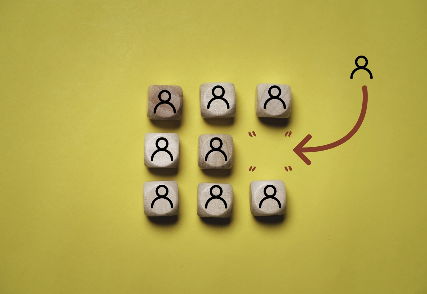 Siete bloques de madera sobre un fondo amarillo. Seis de los bloques están dispuestos en dos filas de tres, cada uno con un icono negro de una persona. El séptimo bloque está separado del grupo a la derecha, con una flecha roja apuntando desde el grupo hacia este bloque solitario.