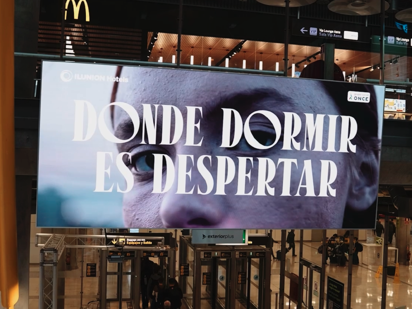 Gran pancarta publicitaria en lo que parece ser un aeropuerto o un espacio público grande. La pancarta presenta un primer plano del rostro de una persona con el texto "DONDE DORMIR ES DESPERTAR" en letras grandes y en negrita. La esquina superior izquierda de la pancarta tiene el logotipo y el nombre "ILUNION Hotels," y la esquina superior derecha tiene un pequeño logotipo con el texto "ONCE." En el fondo, hay letreros que indican direcciones a varias áreas como "VIP Lounge Area" y "Sala Vip Aena." El logotipo de McDonald's también es visible en la esquina superior izquierda de la imagen. La publicidad probablemente está promocionando los hoteles ILUNION con el eslogan "Donde dormir es despertar," sugiriendo una estancia cómoda y refrescante.