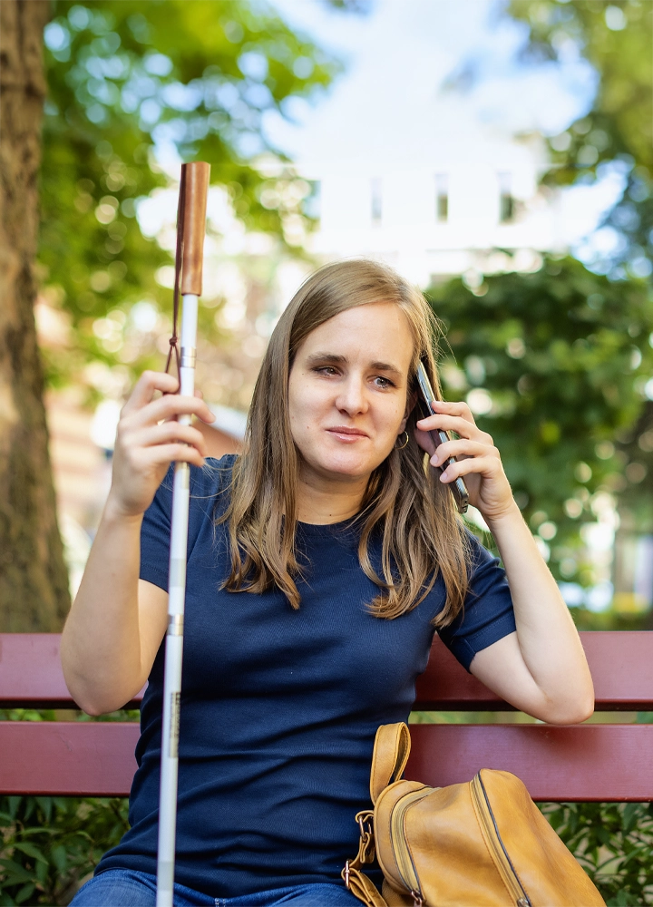 Una mujer joven con cabello castaño está sentada en un banco de parque rojo, sosteniendo un bastón blanco en una mano y hablando por teléfono móvil con la otra. Lleva una camiseta azul y tiene un bolso marrón al lado. El fondo muestra árboles y edificios desenfocados, en un entorno al aire libre con luz natural. La imagen sugiere que la mujer tiene discapacidad visual.