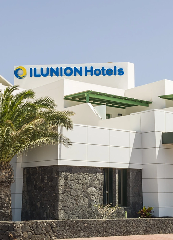Fachada del hotel ILUNION Costa Sal, un establecimiento hotelero ubicado en una zona turística. El edificio, de estilo contemporáneo, cuenta con paredes blancas y detalles en piedra. Una palmera tropical adorna la entrada del hotel, creando un ambiente relajado y acogedor.