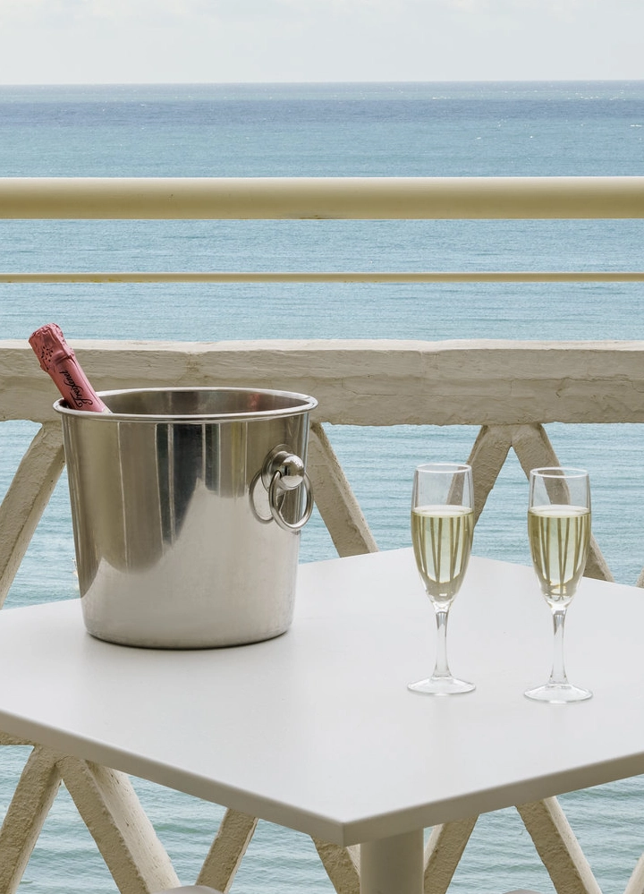 Una mesa pequeña con dos copas de champán y una botella de champagne en un cubo de hielo, ubicada en un balcón con vista al mar.