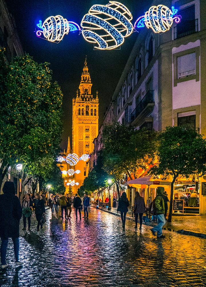 Vista nocturna de una calle peatonal en el centro histórico de Sevilla. La calle está iluminada con farolas y luces navideñas, creando un ambiente festivo. Los edificios antiguos, entre ellos una imponente catedral, se reflejan en el suelo mojado. La gente pasea tranquilamente por la calle, disfrutando del ambiente navideño.