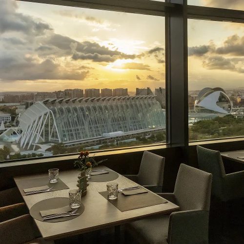restaurante moderno con grandes ventanales, que ofrecen vistas panorámicas a la Ciudad de las Artes y las Ciencias en Valencia. En la mesa hay cliente sentado, un joven trajeado, en la mesa hay copas de cristal y un pequeño jarrón con flores, junto a el podemos leer Umaniko y un código QR. Mientras el cielo nublado en el fondo realza el paisaje urbano.