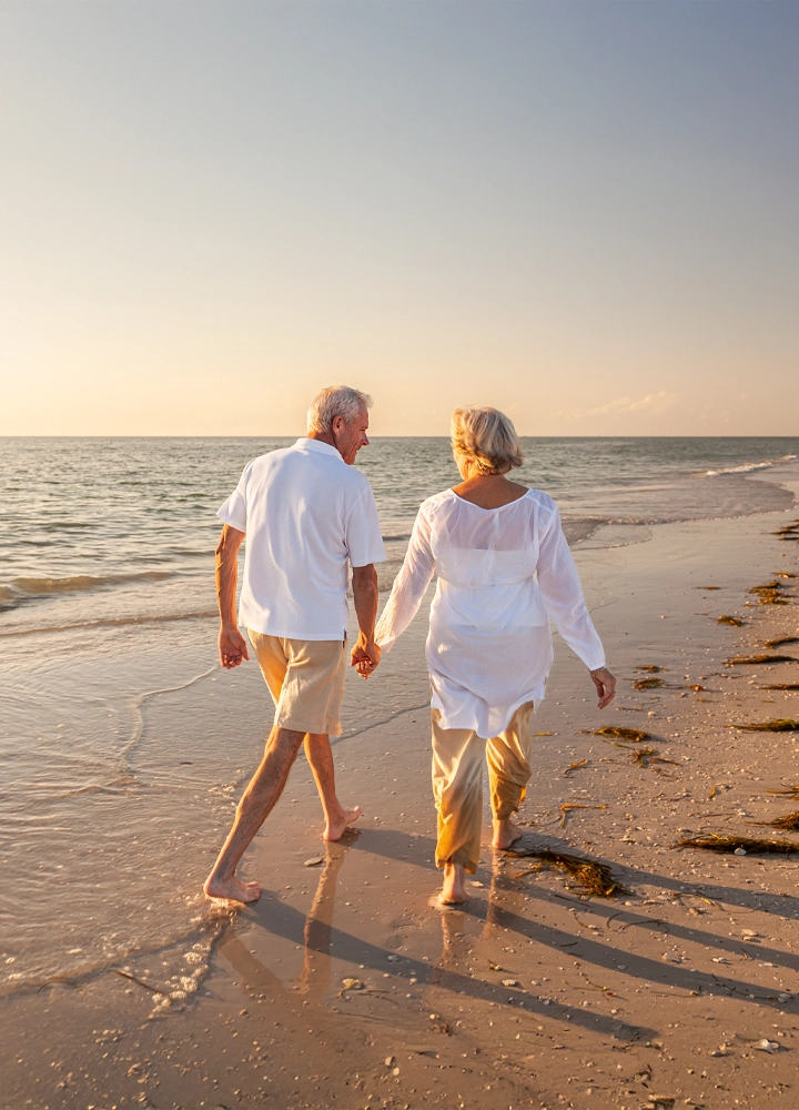 Una pareja de personas mayores camina descalza por la orilla de una playa al atardecer, tomados de la mano. Ambos visten ropa ligera en tonos claros: el hombre lleva una camisa blanca y pantalones cortos beige, mientras que la mujer viste una blusa blanca y pantalones beige. La escena transmite tranquilidad y romanticismo, con el mar a la izquierda y el cielo despejado en tonos cálidos al fondo.