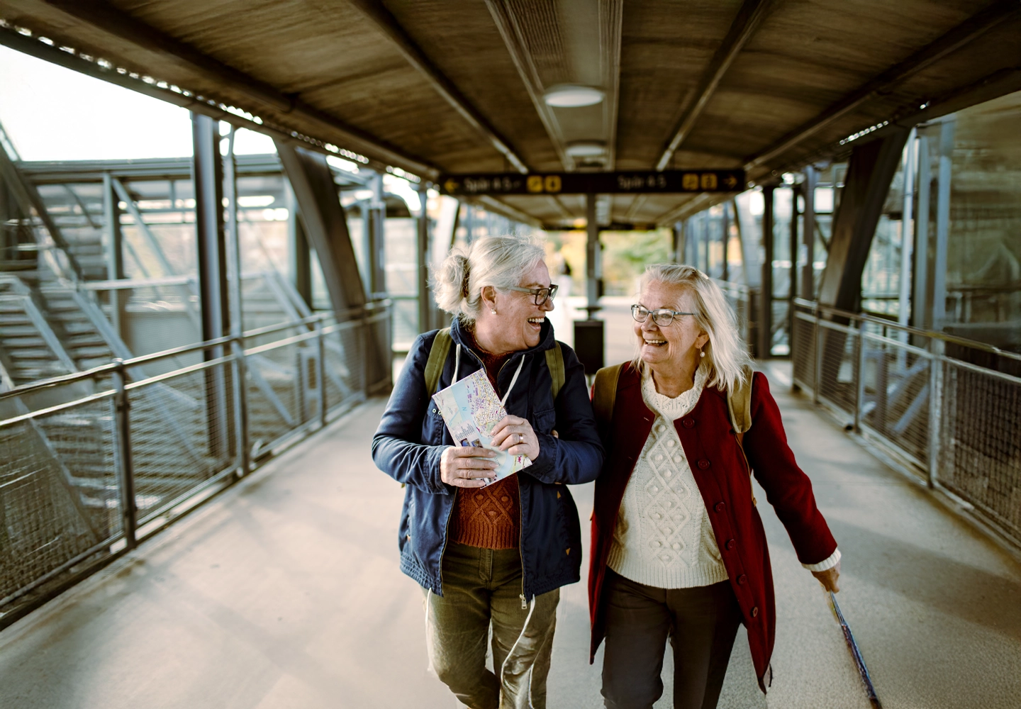 Dos mujeres mayores caminan sonrientes por un pasillo elevado y cubierto en lo que parece ser una estación de transporte. Ambas llevan mochilas y están disfrutando de la conversación. Una de ellas sostiene un mapa en sus manos, mientras que la otra lleva un bastón plegable. La iluminación es cálida, y el entorno combina elementos metálicos y vidrio, indicando un diseño moderno y funcional. La escena refleja alegría y compañerismo durante un viaje.