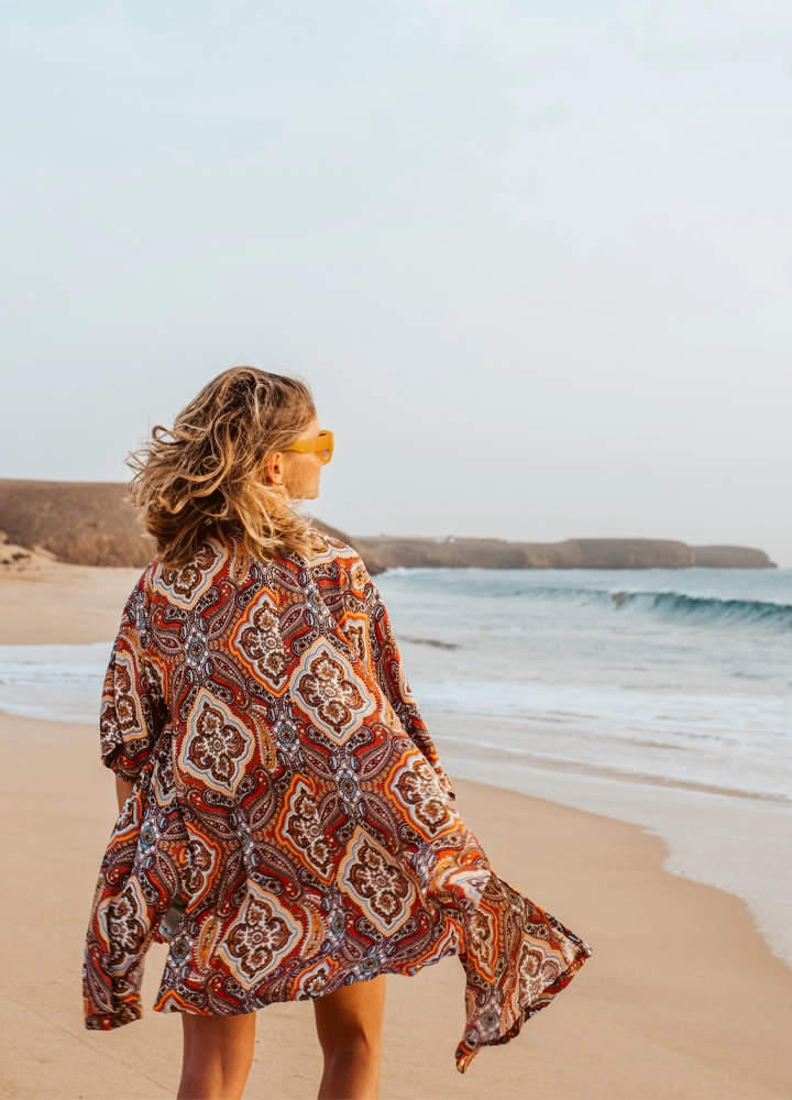 Mujer de cabello rubio y rizado vista de espaldas, caminando por una playa. Lleva una túnica colorida con patrones geométricos en tonos cálidos y gafas de sol amarillas. El mar y las olas están a su derecha, y el paisaje incluye acantilados al fondo bajo un cielo despejado.