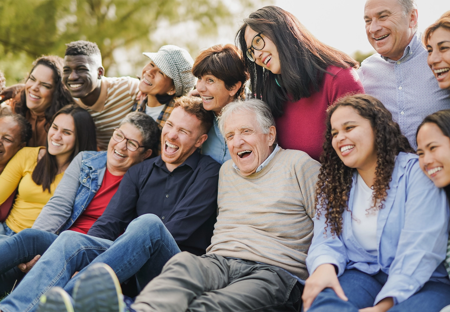 Grupo diverso de personas de diferentes edades y etnias, sentados al aire libre y sonriendo juntos. Transmiten alegría y unión en un entorno natural con árboles de fondo.