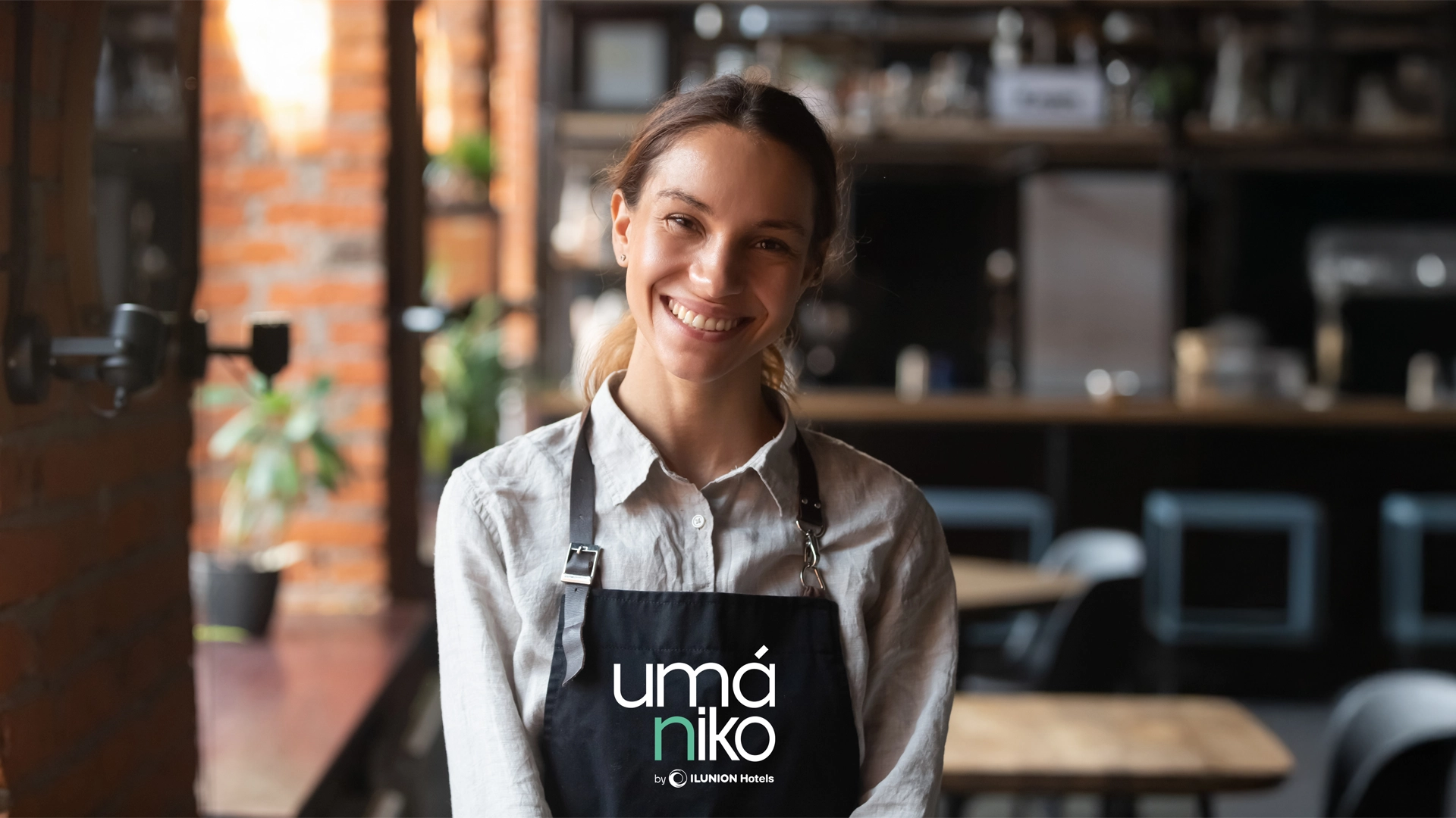 Mujer joven con el cabello recogido en una coleta baja sonríe mientras mira directamente a la cámara. Lleva una camisa blanca y un delantal negro con el logo 'Umániko by ILUNION Hotels'. Se encuentra en el interior de un café o restaurante con un fondo desenfocado de mesas, sillas y estanterías con utensilios.