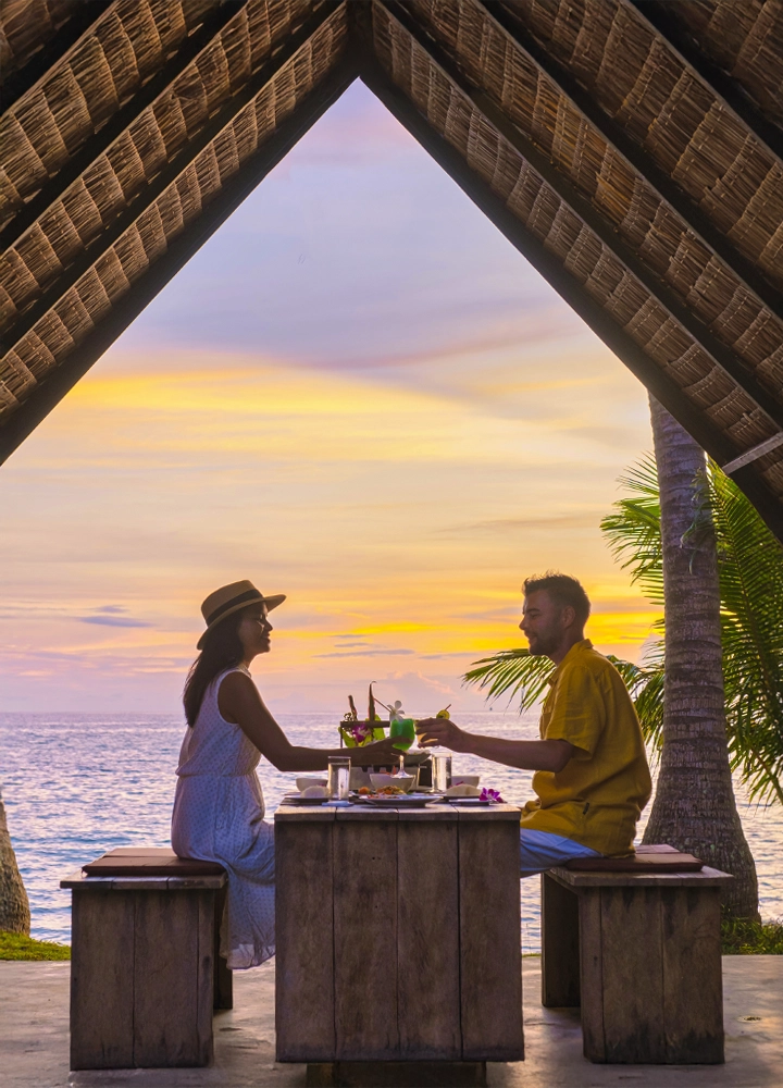 Pareja disfrutando de una cena frente al mar bajo una estructura de techo de palma. La mujer viste un vestido blanco y sombrero, mientras el hombre lleva ropa amarilla. La mesa de madera está decorada con bebidas y platos. De fondo, un atardecer con tonos cálidos y el océano.