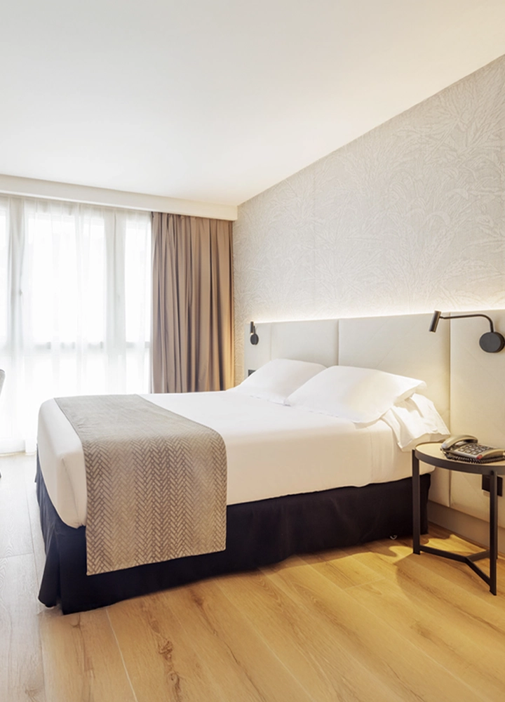 Habitación de hotel moderna con una cama doble vestida con sábanas blancas y manta beige. La pared del cabecero tiene un diseño texturizado, con luces empotradas y lámparas de lectura. Hay una mesa auxiliar con teléfono y el suelo es de madera clara. La habitación recibe luz natural a través de una gran ventana con cortinas.