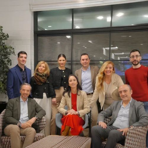 Grupo de nueve personas posando juntas en el interior de una sala moderna con decoración corporativa. Tres personas están sentadas en sillas frente a dos mesas de diseño en forma de barril, mientras que las otras seis están de pie detrás. La sala cuenta con un suelo de madera clara, una pared con ventanales, y plantas decorativas a la izquierda.