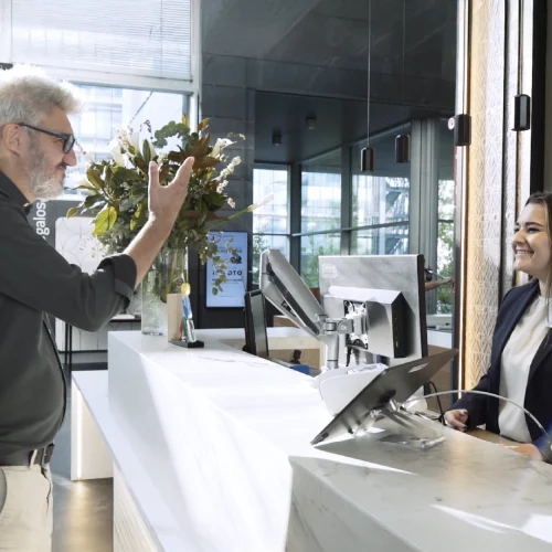 Hombre con cabello canoso, gafas y camisa oscura interactuando con una recepcionista de hotel que viste un traje formal y sonríe detrás de un mostrador de mármol blanco. En el fondo, un gran ventanal deja entrar luz natural, con plantas decorativas y pantallas en la recepción.