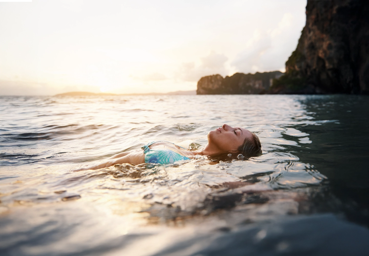 Mujer flotando de espaldas en el agua durante el atardecer, con una expresión relajada y los ojos cerrados. Lleva un bikini con estampado tropical. El paisaje incluye acantilados oscuros al fondo y un cielo cálido con tonos dorados reflejados en el agua.