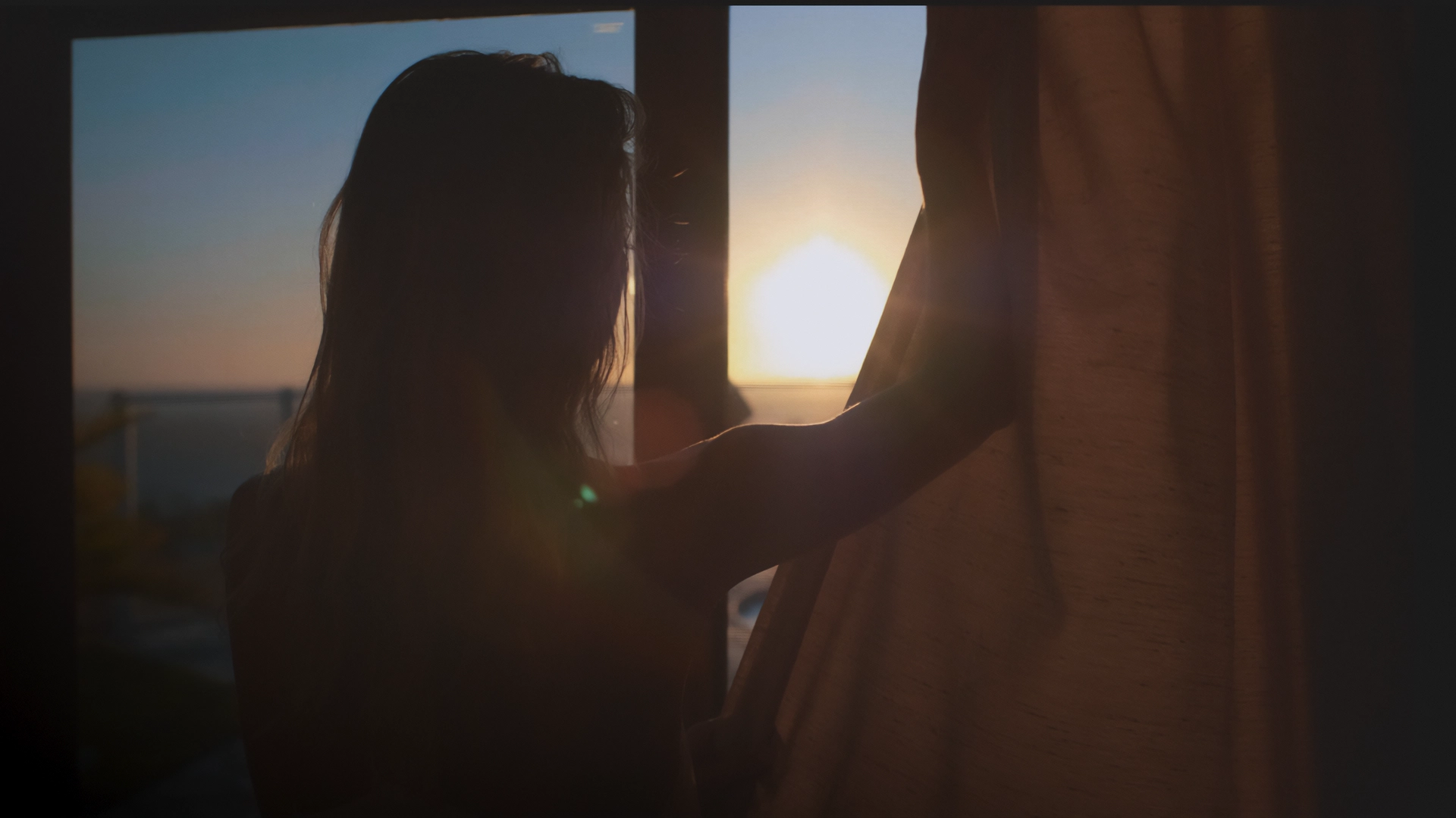 Silueta de una mujer abriendo una cortina frente a una ventana al amanecer o atardecer. La luz cálida del sol entra por la ventana, iluminando suavemente el espacio y creando un ambiente tranquilo