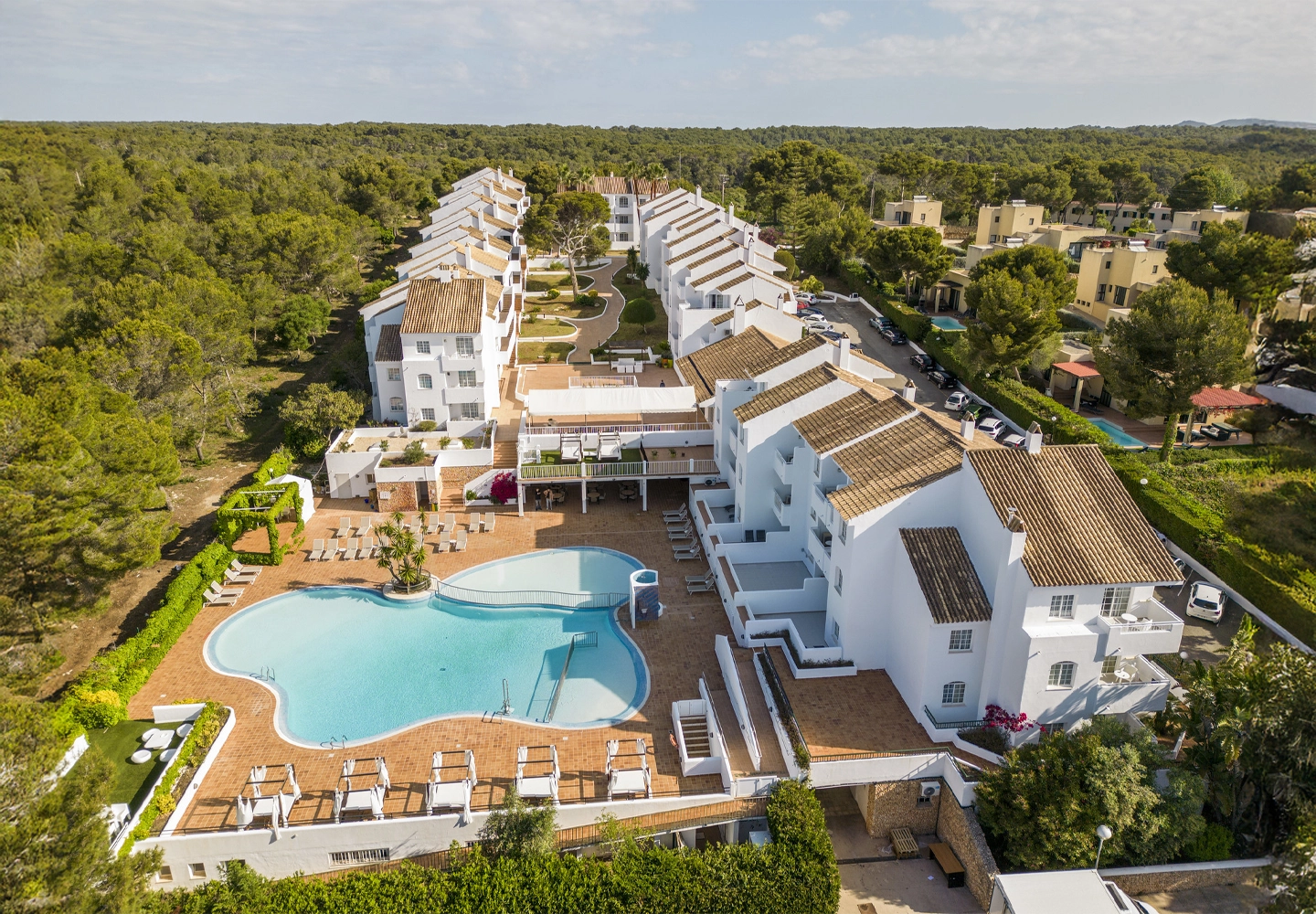 Vista aérea de un complejo turístico con edificios de fachadas blancas y techos de tejas. En primer plano, una piscina de forma orgánica rodeada de tumbonas y sombrillas. El complejo está ubicado junto a una extensa área boscosa y áreas residenciales.