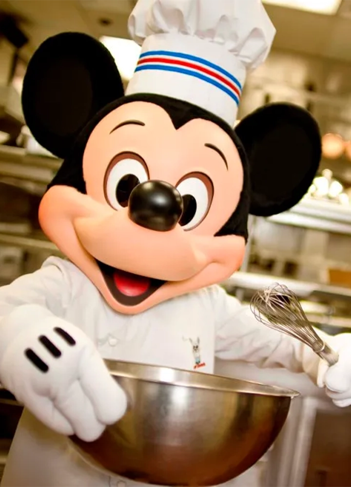 Un personaje disfrazado de ratón, con características de dibujos animados, vestido de chef. Lleva un gorro de cocina blanco con una banda tricolor y un uniforme blanco de chef. Sostiene un bol metálico en una mano y un batidor en la otra, mientras sonríe ampliamente en una cocina profesional.