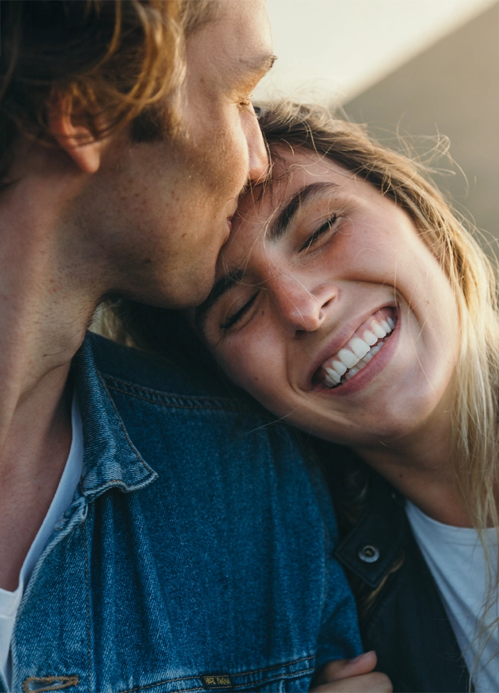 Primer plano de una pareja mostrando un momento de cariño y felicidad. La mujer sonríe ampliamente mientras apoya su cabeza en el hombro del hombre, quien le da un beso en la frente. Ambos transmiten cercanía y alegría, con el hombre vistiendo una chaqueta vaquera y un fondo desenfocado que aporta calidez a la escena.