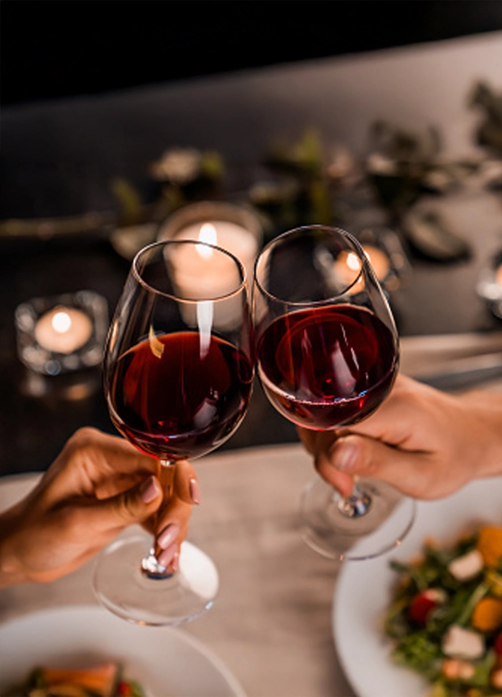 Primer plano de dos manos brindando con copas de vino tinto sobre una mesa puesta para una cena.