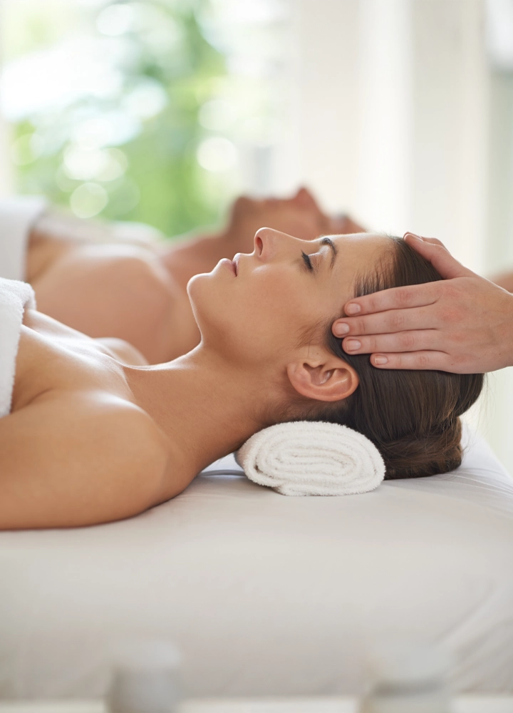 Primer plano de una mujer recibiendo un masaje relajante en la cabeza, tumbada sobre una camilla con una toalla enrollada bajo su cuello. En el fondo, desenfocado, se observa otra persona también tumbada en una camilla. El entorno es luminoso y sereno, con un fondo de vegetación que refuerza la sensación de bienestar y tranquilidad.