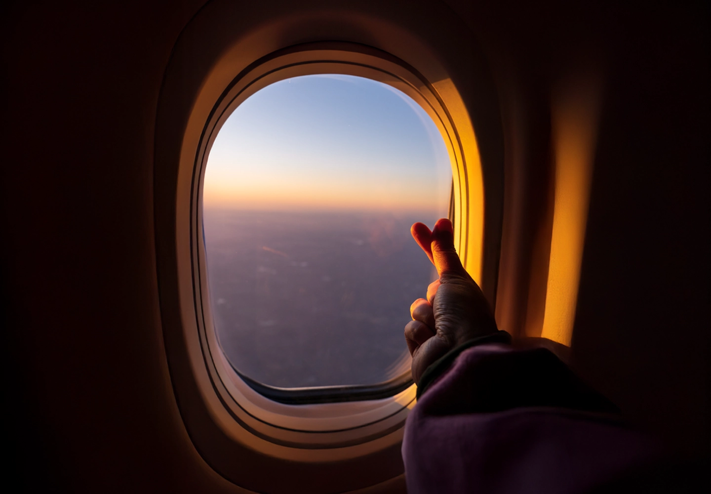 Primer plano de una mano formando un corazón con los dedos, con el fondo de una ventana de avión y el cielo anaranjado al atardecer.
