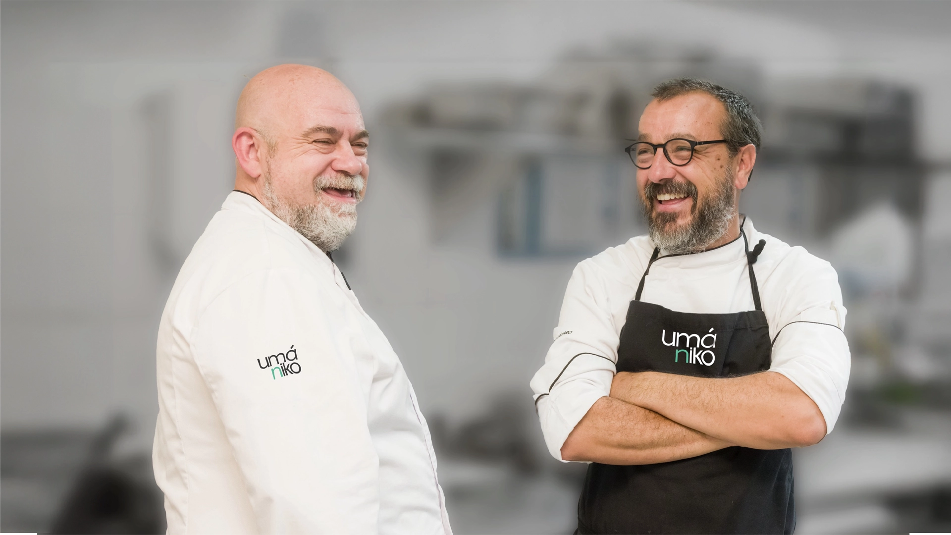 Dos chefs sonrientes posan en una cocina. Ambos visten chaquetas blancas de chef con el logo &#039;umániko&#039;. Uno de ellos lleva también un delantal negro. La escena refleja camaradería y profesionalismo en el entorno culinario.