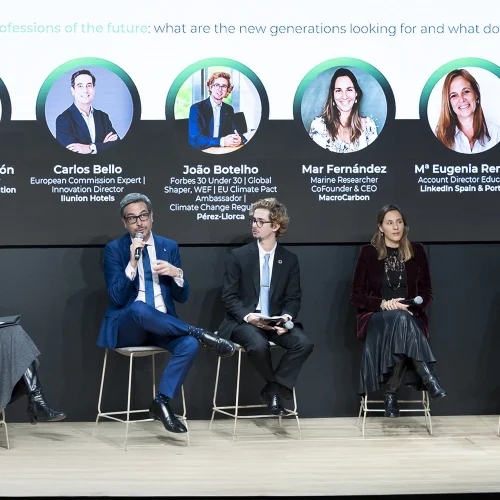 Evento profesional sobre sostenibilidad y profesiones del futuro. En el escenario, seis panelistas están sentados en taburetes, incluidos profesionales destacados como Concepción Galdón, Carlos Bello, João Botelho, Mar Fernández, Mª Eugenia Renaldi y Belén Santos, cuyos nombres y cargos están proyectados en la pantalla detrás de ellos. Cada panelista está vestido de manera formal. Carlos Bello, con traje azul, está hablando al micrófono mientras los demás escuchan con atención. Una moderadora está sentada a la izquierda, sosteniendo una tablet. El entorno es moderno, con un fondo oscuro y un diseño minimalista.