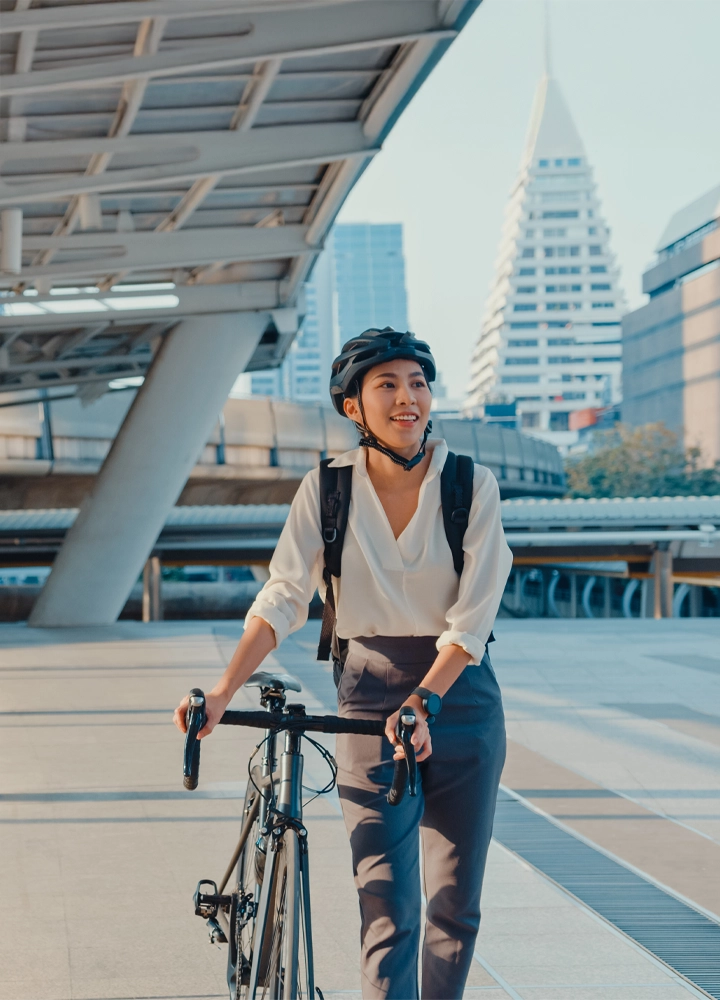 Mujer joven con casco caminando junto a su bicicleta en una ciudad moderna con edificios altos de fondo