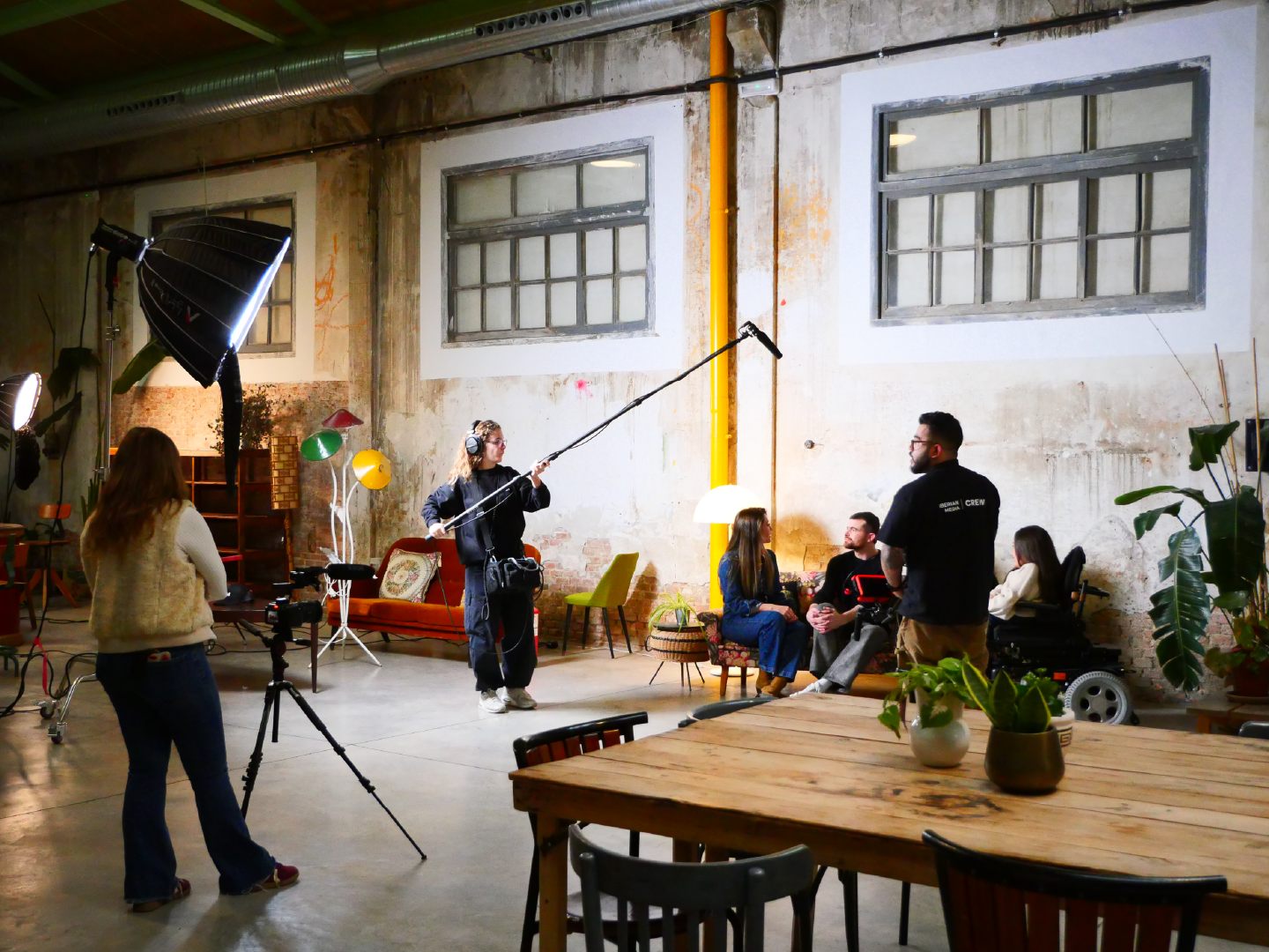 Equipo de producción graba una escena en un estudio de aspecto industrial con cámaras, micrófonos y luces. Varias personas están reunidas en el fondo conversando.