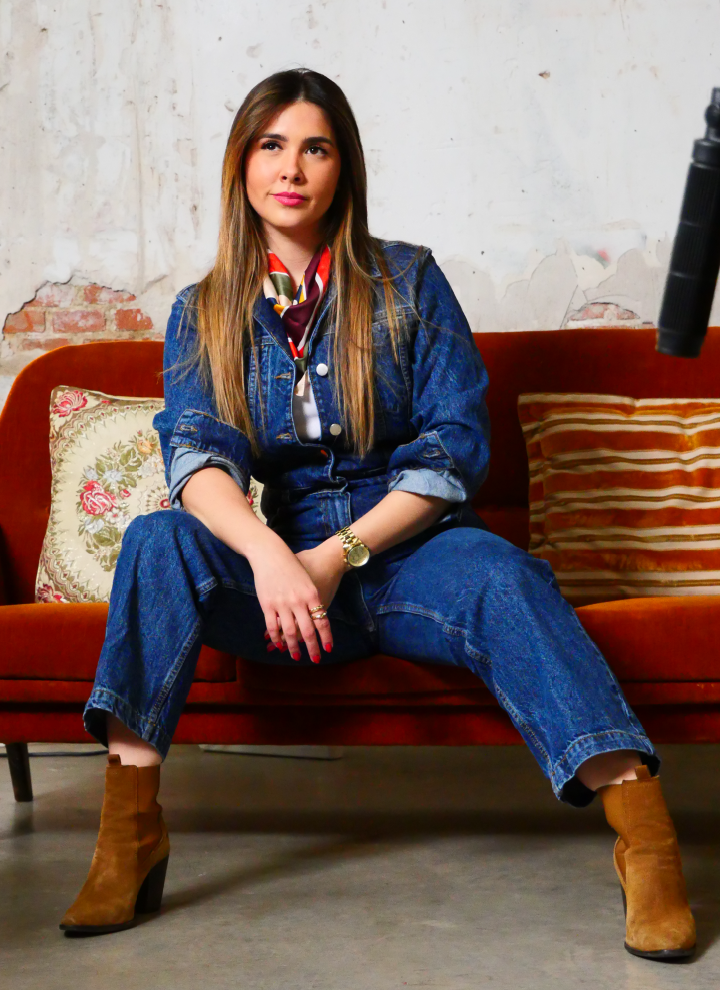 Una persona sentada en un sofá rojo de aspecto vintage. La persona lleva un conjunto de mezclilla, que incluye una chaqueta de mezclilla y unos jeans, combinado con botines marrones. También lleva un reloj en la muñeca izquierda y varios anillos en los dedos. En el fondo, se ve una pared desgastada con partes de ladrillos expuestos, lo que refuerza la estética vintage