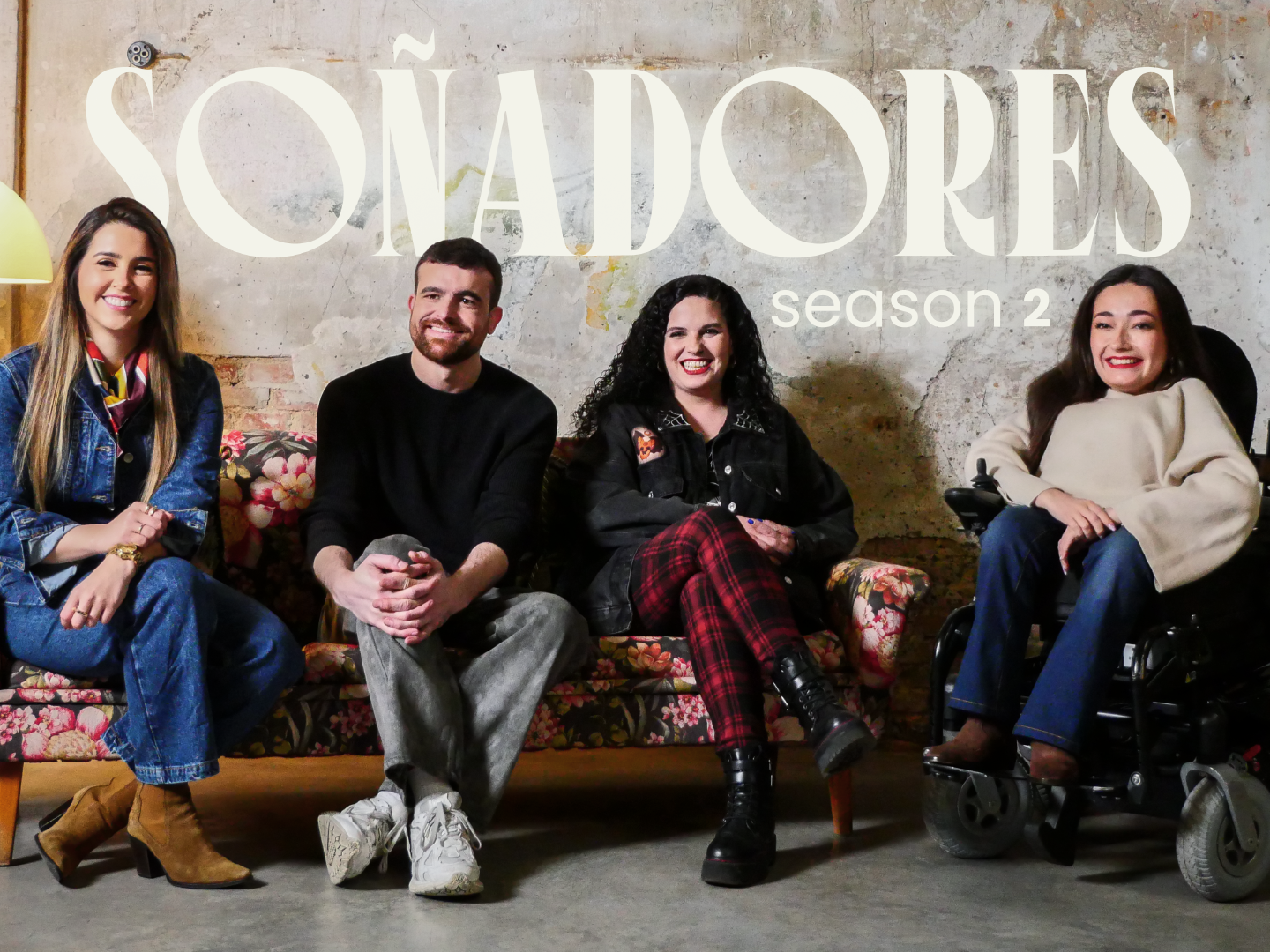 Cuatro personas sonrientes están sentadas en un sofá vintage con estampado floral frente a una pared de cemento con textura envejecida. Sobre ellos, en grandes letras blancas, se lee “SOÑADORES season 2”. A la izquierda, una mujer con chaqueta vaquera, pañuelo de colores y botas marrones. A su lado, un hombre con jersey negro y pantalones grises, con las manos entrelazadas. Junto a él, una mujer de cabello rizado y chaqueta negra con tachuelas, vistiendo pantalón de cuadros y botas negras. A la derecha, una mujer con blusa beige y pantalón azul, sentada en una silla de ruedas eléctrica, con una gran sonrisa. La imagen refleja un ambiente acogedor y auténtico, con un estilo industrial y bohemio.
