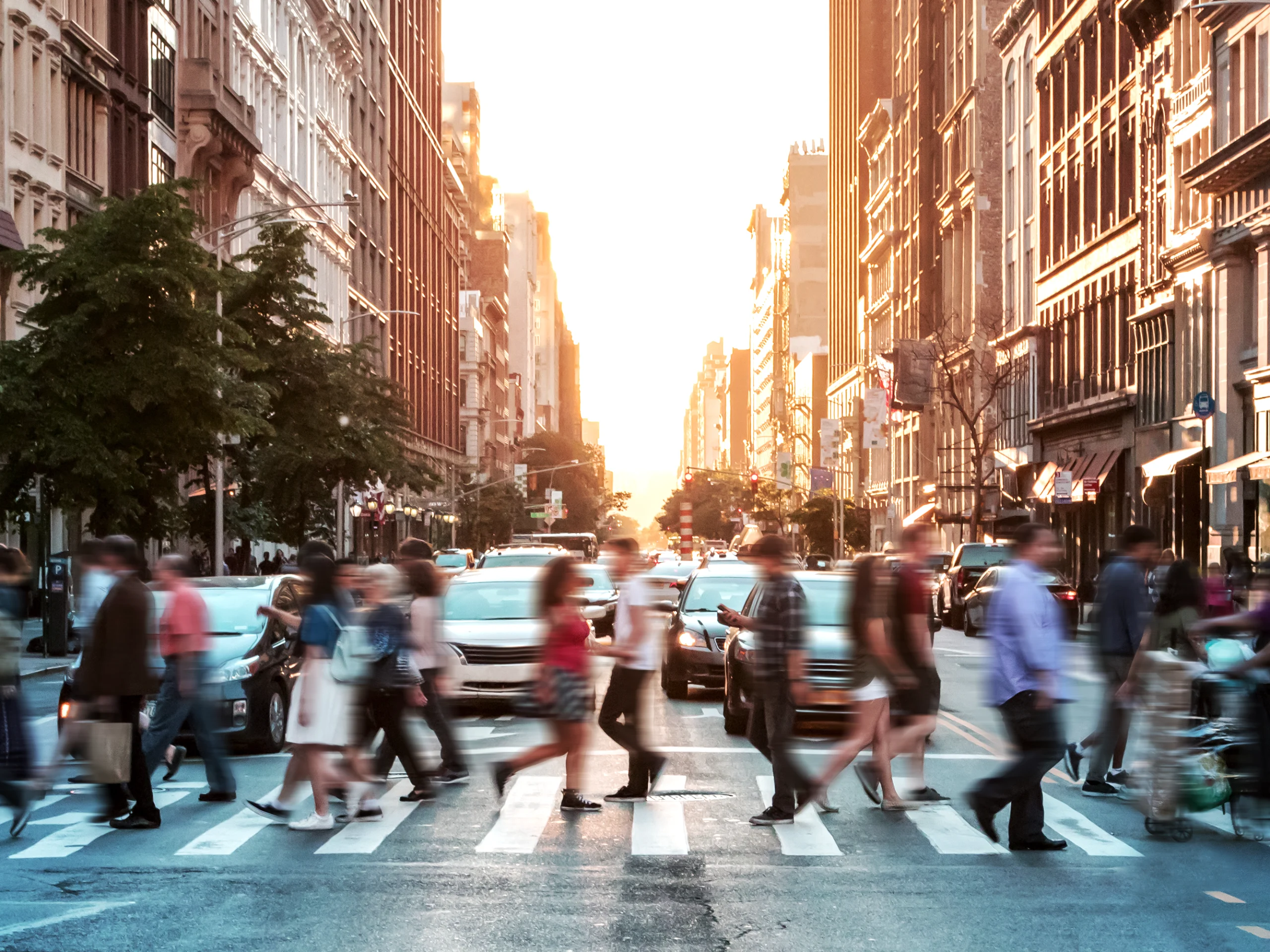Cruce peatonal concurrido en una avenida urbana al atardecer, con edificios altos a ambos lados. Personas caminando rápidamente en diferentes direcciones mientras los vehículos esperan en el cruce. La luz dorada del sol ilumina el fondo, creando un contraste entre el movimiento de la gente y la arquitectura estática.