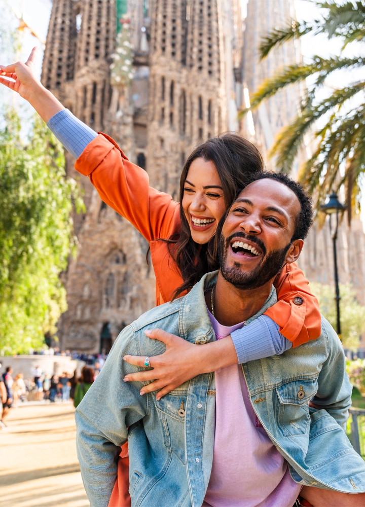 Pareja feliz riendo mientras él la lleva a cuestas frente a la Sagrada Familia en Barcelona.