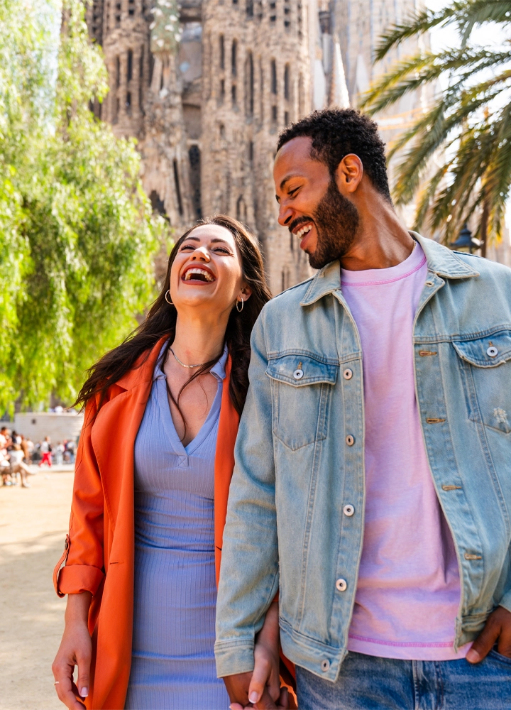 Pareja sonriendo mientras camina tomada de la mano frente a la Sagrada Familia en Barcelona.