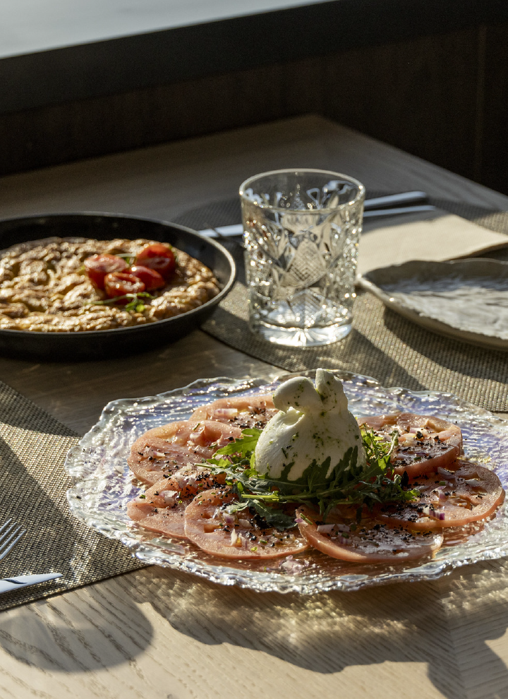 Un plato elegante servido en una mesa con luz natural. En primer plano, un carpaccio de tomate con burrata en el centro, decorado con rúcula y especias, sobre un plato de cristal con diseño detallado. En el fondo, un plato de berenjenas asadas con tomates cherry y un vaso tallado con agua. La escena transmite una atmósfera sofisticada y acogedora, con una cuidada presentación de la comida.