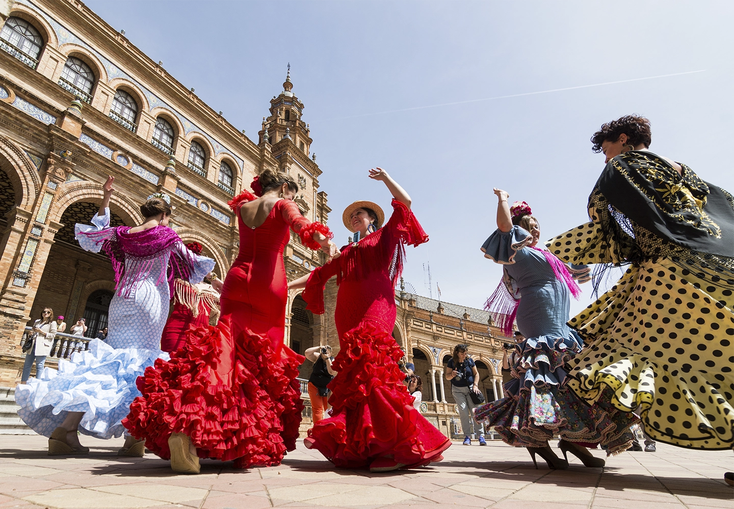 Un grupo de mujeres vestidas con trajes tradicionales de flamenco bailan en una plaza. Los vestidos son coloridos, con volantes y lunares, destacando los colores rojo, azul y amarillo. Al fondo se observa un edificio histórico con arcos y una torre.