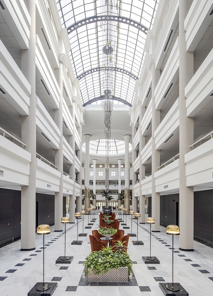 Amplio vestíbulo interior con un techo de vidrio arqueado y columnas altas a ambos lados. En el centro hay una fila de sofás marrones y mesas con plantas verdes, flanqueadas por lámparas doradas. El suelo es de mármol blanco con detalles en negro.